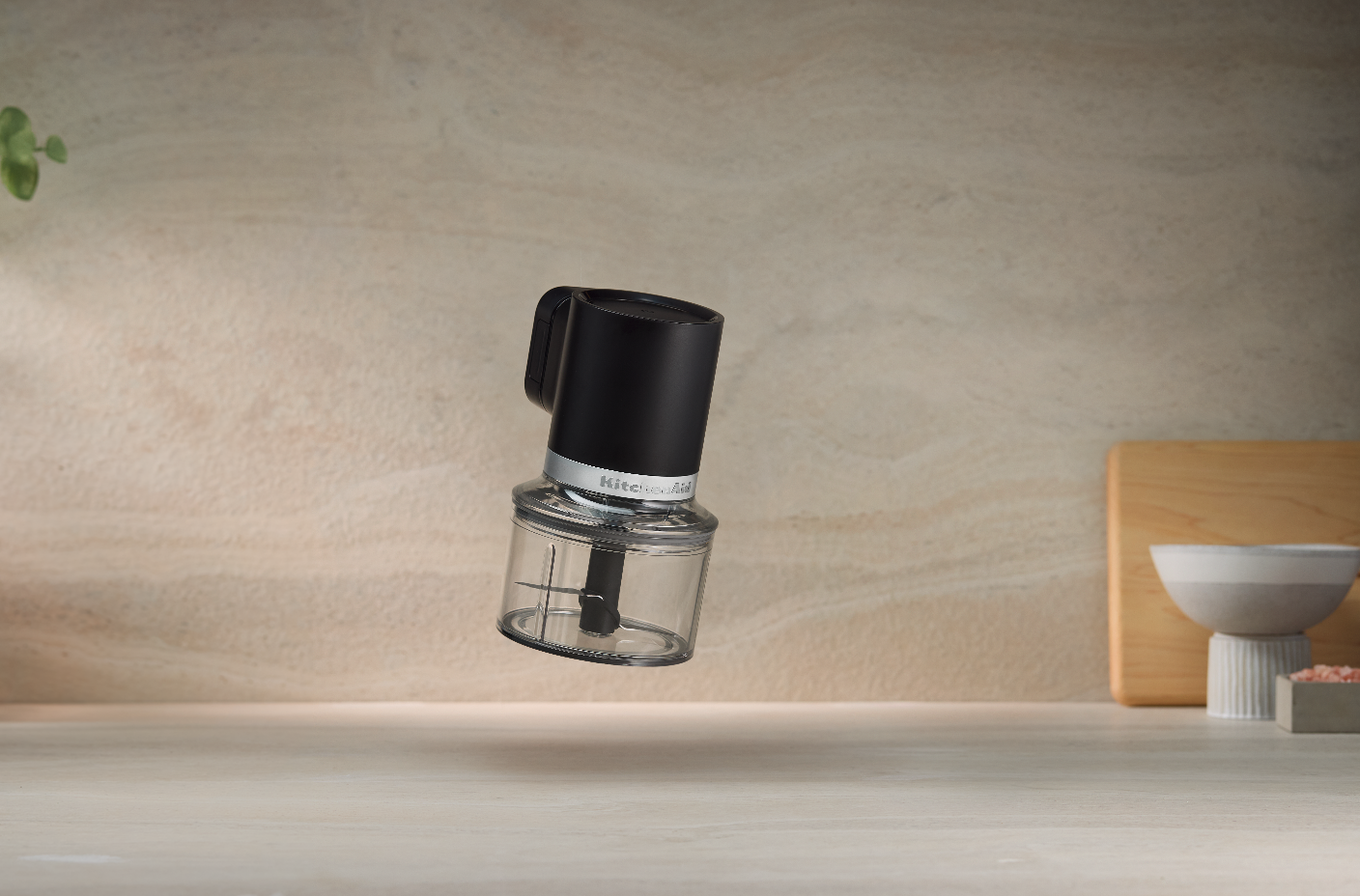 Dark-finish KitchenAid Go cordless top-down chopper poised above its clear bowl, with the central blade assembly visible.