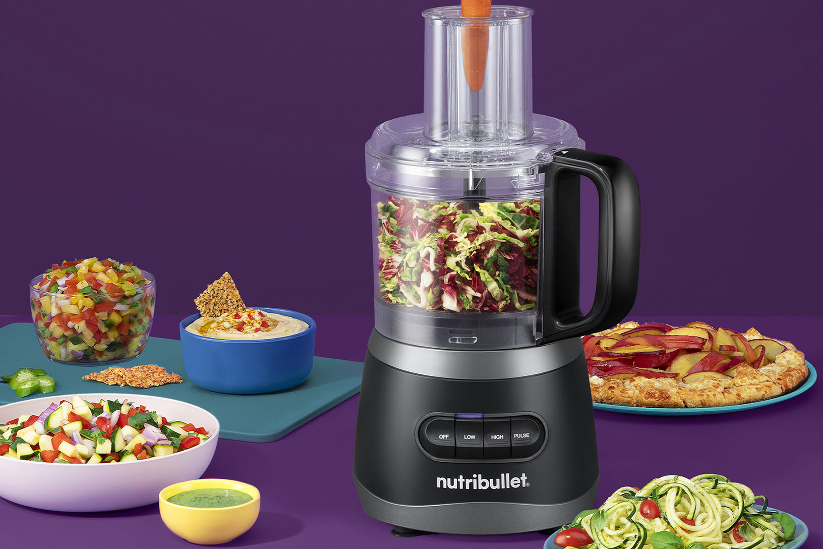 Black-and-silver nutribullet standalone food processor with clear work bowl, feed chute, pusher and OFF/LOW/HIGH/PULSE controls, shown with prepared food.